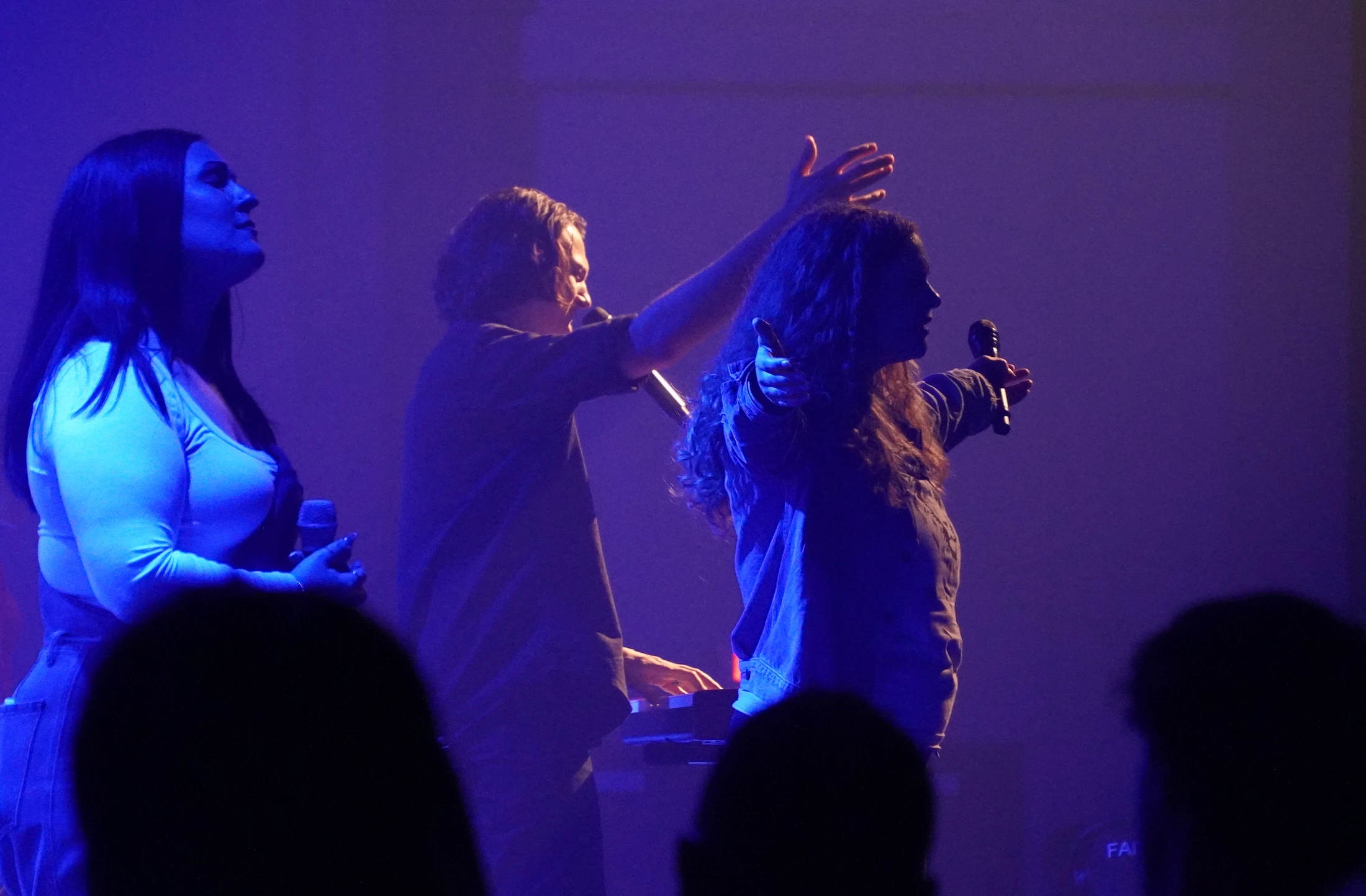Three musicians on a dimly lit stage bathed in purple and blue light. Two women sing in profile while a man in the center plays a keyboard.